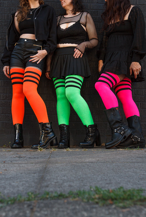 3 people wearing Electric Shock thigh highs in neon green, neon orange, and neon pink, all with black stripes.