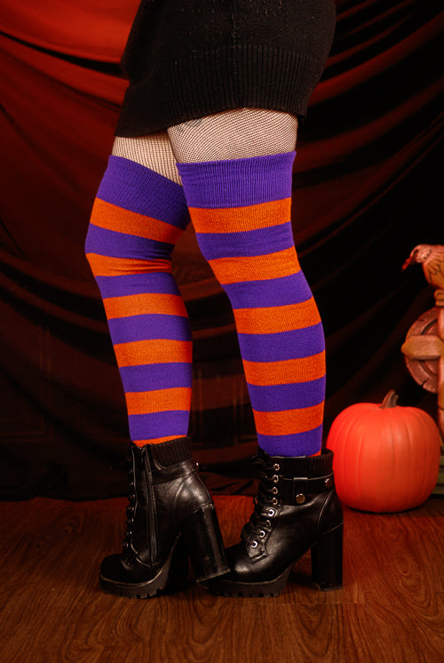 Person wearing a black sweater with, black skirt, and striped orange and purple thigh highs against a dark background.