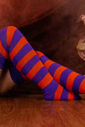 Person wearing a black sweater with, black skirt, and striped orange and purple thigh high socks against a dark background.