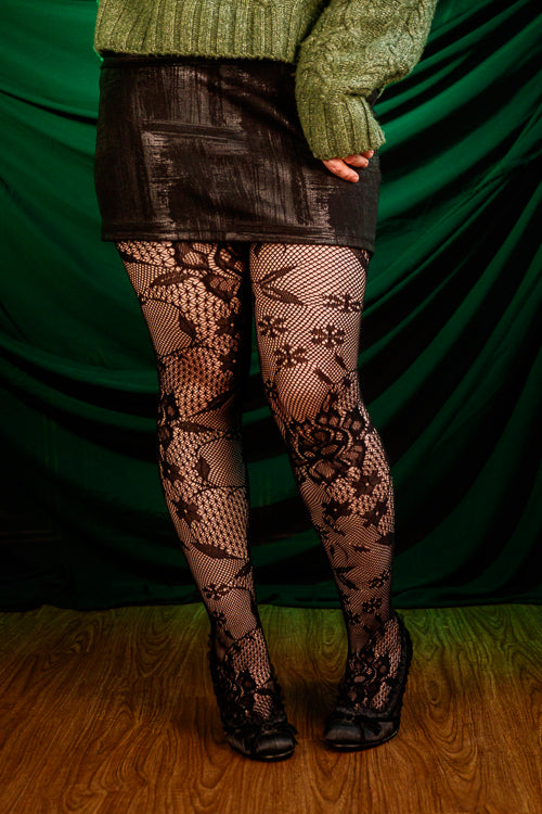 Person wearing a green sweater, black patterned floral tights, and a black skirt against a green curtain.