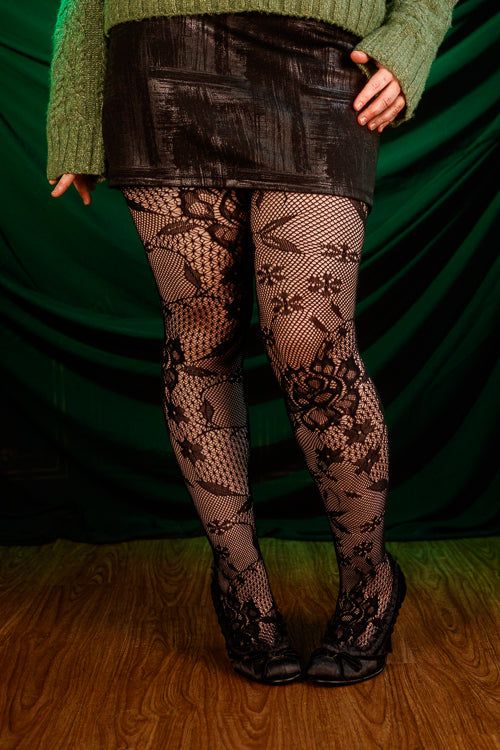 Person wearing a green sweater, black patterned floral tights, and a black skirt against a green curtain.