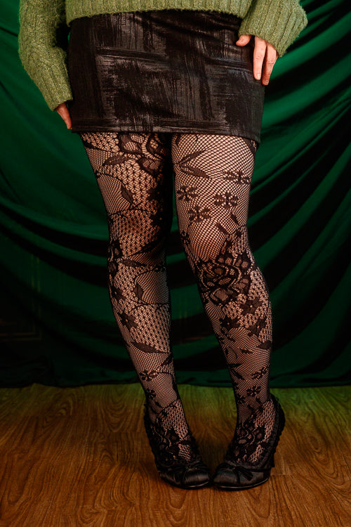 Person wearing a green sweater, black patterned floral tights, and a black skirt against a green curtain.