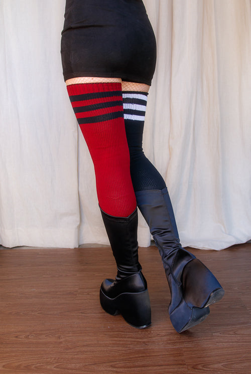 A person wearing one black thigh high with white top-stripes, and one red thigh high with black top stripes