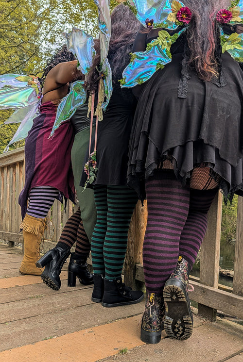 A group of four people wearing different colors of Faery stripes and fairy wings, standing on a wooden bridge.