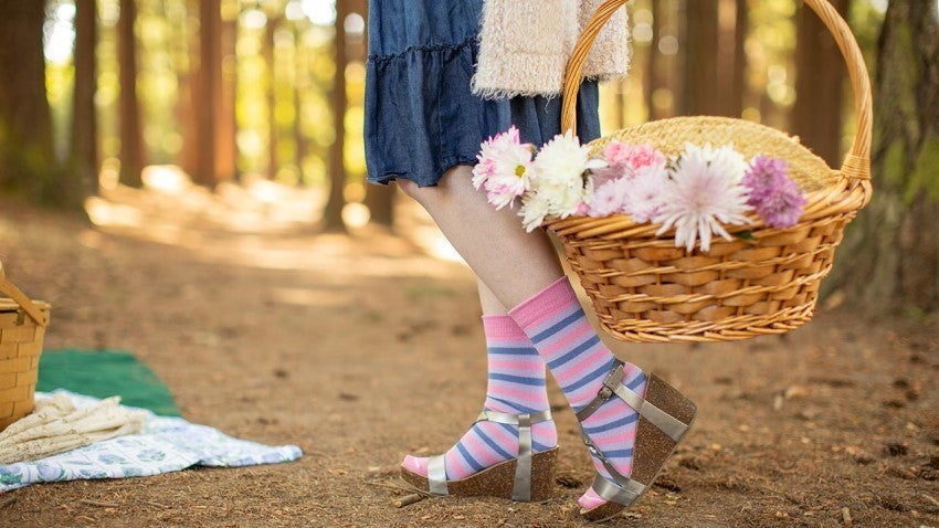 Person wearing Pastel Pride Crew Socks - Bisexual Colorway, holding a wicker basket filled with flowers in a sunny pine forest.