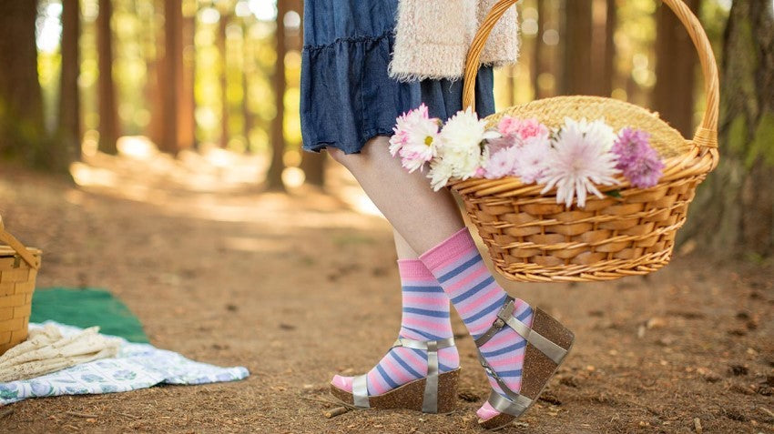Person wearing Pastel Pride Crew Socks - Bisexual Colorway, holding a wicker basket filled with flowers in a sunny pine forest.