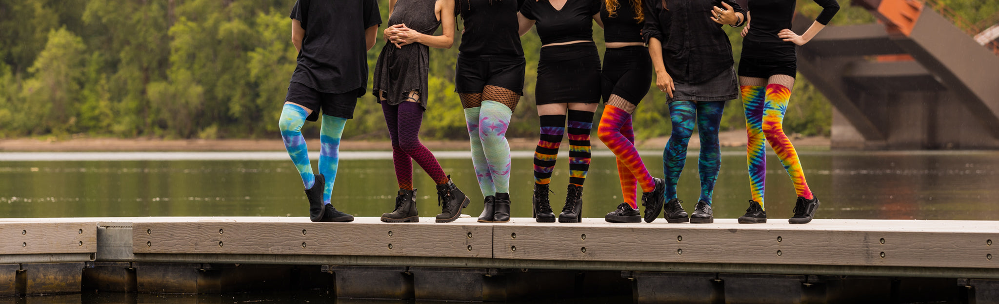 Seven people, each wearing a different pair of tie-dyed thigh high socks. They stand along a dock with a placid river and greenery in the background.