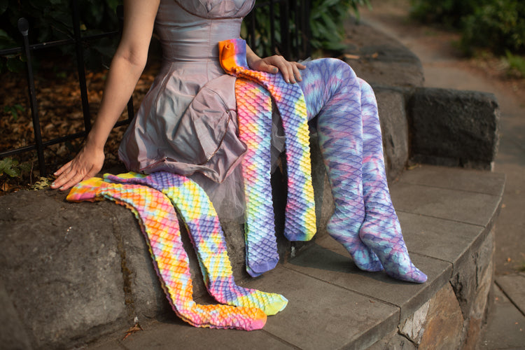 A person wearing long socks tie-dyed with a diamond pattern of pastel blues, purples, and pinks sitting on a raised stone bench. She displays several other pairs of the same sock style in different pastel tie dye designs on the ledge next to her.