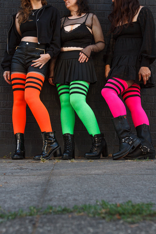 3 people wearing Electric Shock thigh highs in  neon green, neon orange, and neon pink, all with black stripes.