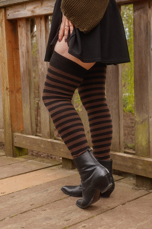 A person wearing black and dark brown fairy striped thigh-high socks, in boots on a wooden deck.