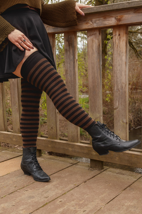 A person wearing black and dark brown fairy striped thigh-high socks, in boots on a wooden deck.