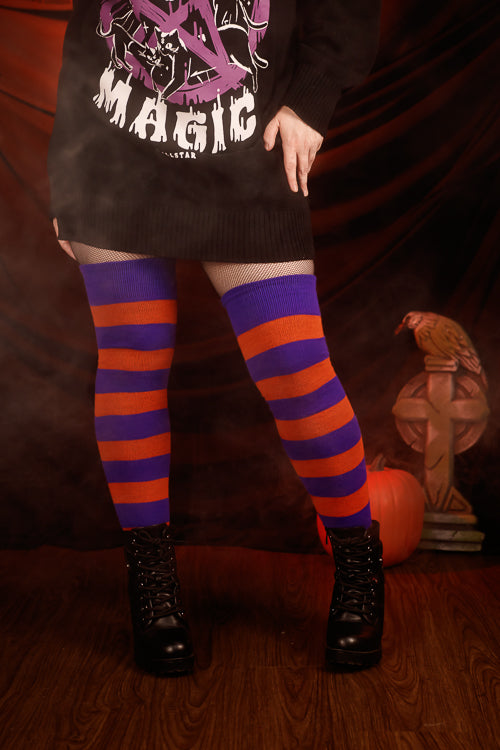 Person wearing a black sweater with purple and white design, black skirt, and striped orange and purple thigh highs against a dark background.