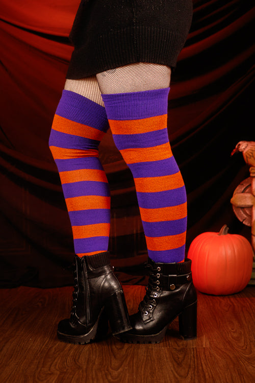 Person wearing a black sweater with, black skirt, and striped orange and purple thigh highs against a dark background.