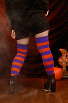 Person wearing a black sweater with, black skirt, and striped orange and purple thigh highs against a dark background.