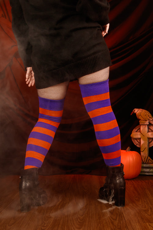 Person wearing a black sweater with, black skirt, and striped orange and purple thigh highs against a dark background.