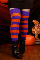 Person wearing a black sweater with, black skirt, and striped orange and purple thigh highs against a dark background.