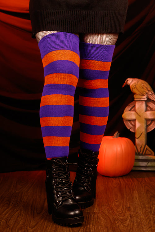 Person wearing a black sweater with, black skirt, and striped orange and purple thigh highs against a dark background.