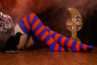 Person wearing a black sweater with, black skirt, and striped orange and purple thigh high socks against a dark background.