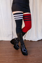 A person wearing one black thigh high with white top-stripes, and one red thigh high with black top stripes