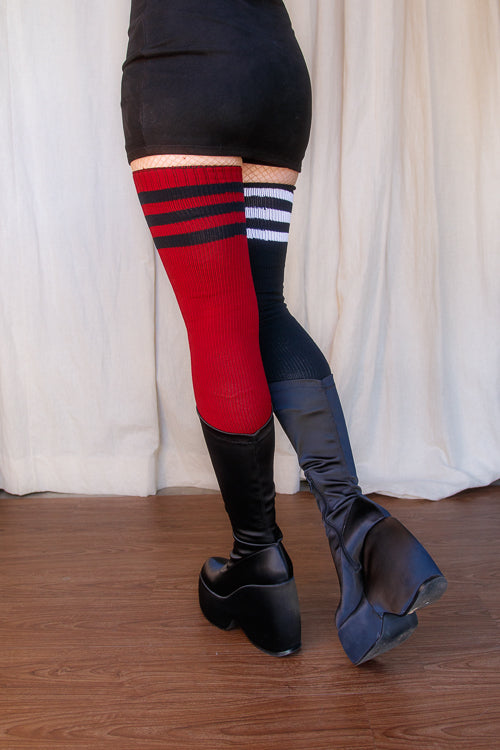A person wearing one black thigh high with white top-stripes, and one red thigh high with black top stripes