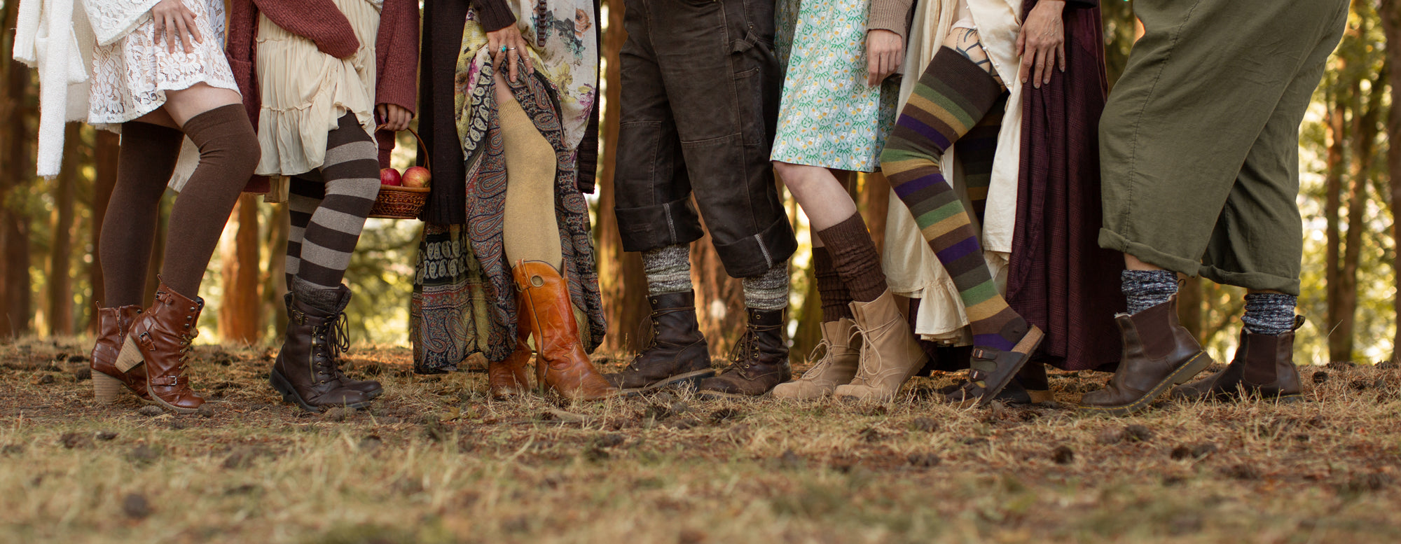 Seven people wearing a variety of tall and knee length socks with a color palette centered on browns, neutrals, and autumn colors. They stand in a sunny clearing of trees with light brown grasses and pinecones underfoot.